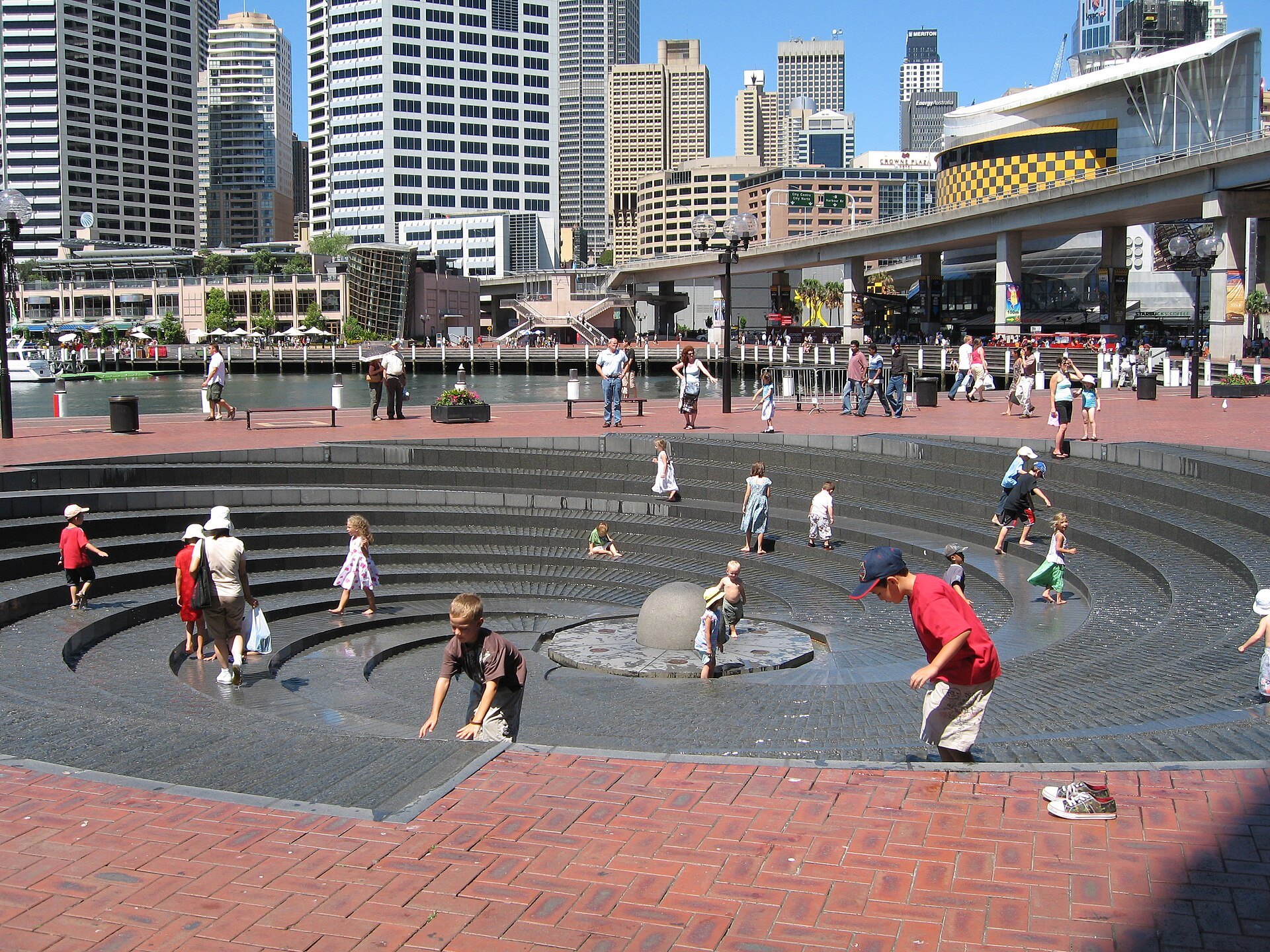 The paved plaza at Darling Harbour with people walking and the city towers behind