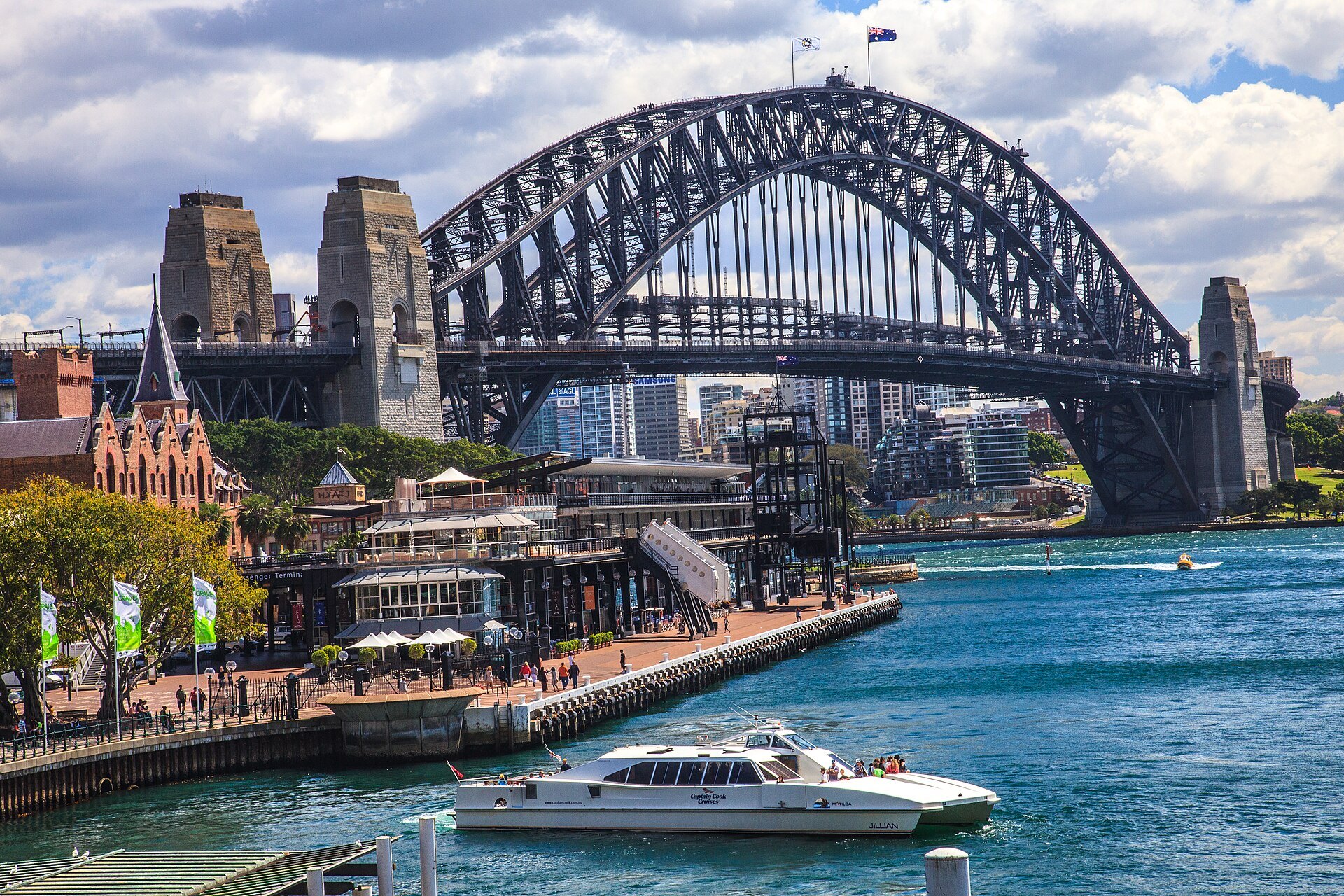 A Sydney ferry at Circular Quay with the Harbour Bridge behind it, the sandstone buildings of the quay on the left and the harbour bright behind