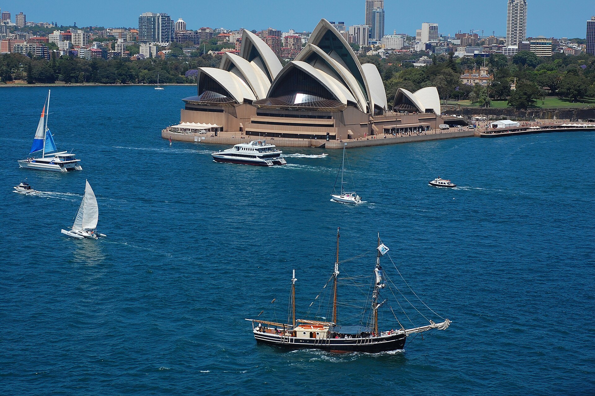 The Opera House from the water with a tall ship and yachts sailing past, the city behind